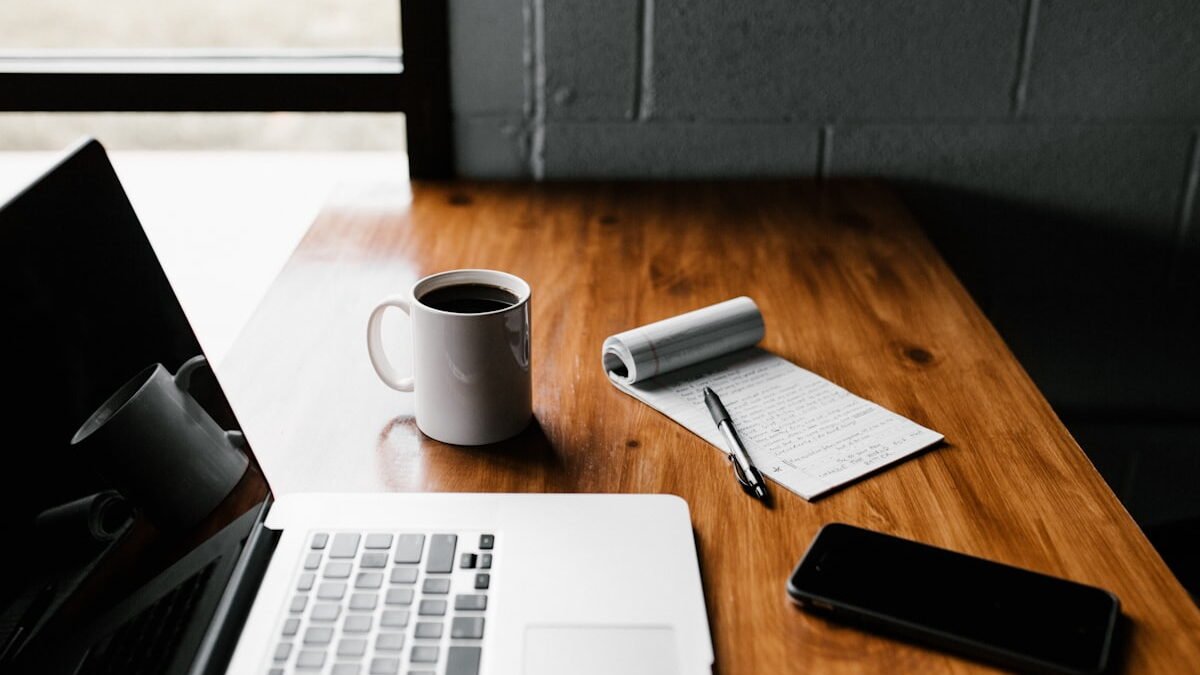 A laptop, a cup of black coffee and a handwritten notebook on a wooden desk by a window